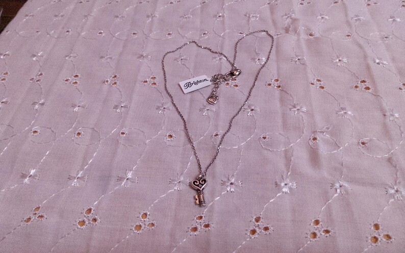 Op de afbeelding: Een zilverkleurige ketting met een sleutelhanger. De sleutelhanger heeft een hartvormige bovenkant en een klein sleutelontwerp. De ketting heeft een label met de tekst "Brighton". De ketting wordt getoond op een lichtroze, geborduurde stof.