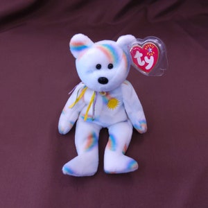 May include: A white rainbow-patterned beanie baby bear with a yellow sun and purple ribbon. The bear has a red heart-shaped tag with the text "Beanie Baby" and the Ty logo.