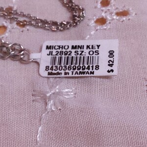 Op de afbeelding: Een zilverkleurige ketting met een label met de tekst "MICRO MNI KEY JL2892 SZ: OS" en een barcode. Op het label staat ook "Made in TAIWAN". De ketting ligt op een lichtroze, geborduurde stof.