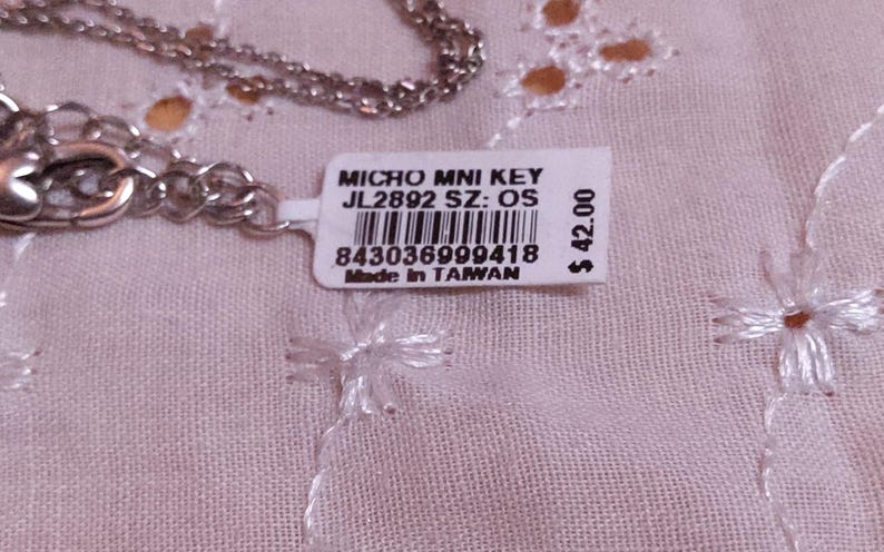 Op de afbeelding: Een zilverkleurige ketting met een label met de tekst "MICRO MNI KEY JL2892 SZ: OS" en een barcode. Op het label staat ook "Made in TAIWAN". De ketting ligt op een lichtroze, geborduurde stof.