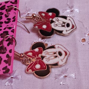 Pu&ograve; includere: Un paio di orecchini pendenti di Minnie Mouse. Ogni orecchino presenta un viso bianco con dettagli neri, un fiocco rosso a pois bianchi e orecchie nere. Gli orecchini sono dorati e hanno una chiusura a gancio.