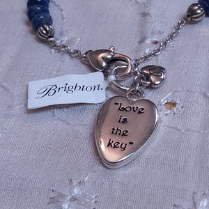 Puede incluir: Collar plateado con un colgante en forma de coraz&oacute;n que dice "Love is the key". El collar tiene una secci&oacute;n de cuentas azules y una etiqueta "Brighton".