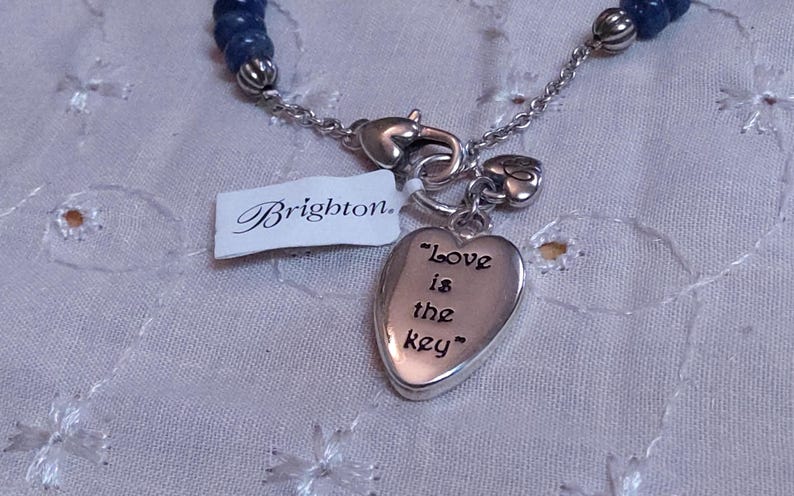 Puede incluir: Collar plateado con un colgante en forma de coraz&oacute;n que dice "Love is the key". El collar tiene una secci&oacute;n de cuentas azules y una etiqueta "Brighton".