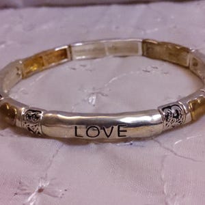 May include: A silver bracelet with gold accents and the word "LOVE" engraved on a central bar. The bracelet is made of multiple small beads with decorative designs.