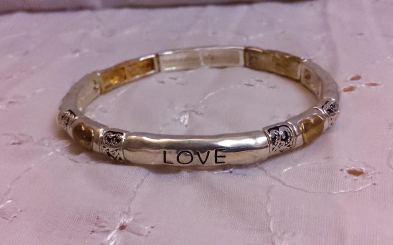 May include: A silver bracelet with gold accents and the word "LOVE" engraved on a central bar. The bracelet is made of multiple small beads with decorative designs.