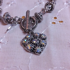 May include: A silver bracelet with a heart-shaped charm. The charm is decorated with iridescent rhinestones. The bracelet has a toggle clasp.