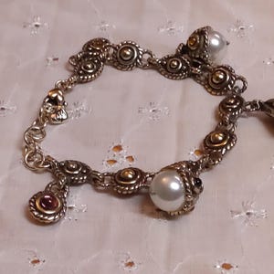 May include: Silver-toned bracelet with gold accents, featuring round and oval links. The bracelet includes pearl and gemstone charms. The design incorporates ornate detailing and a vintage aesthetic, suitable for jewelry enthusiasts.
