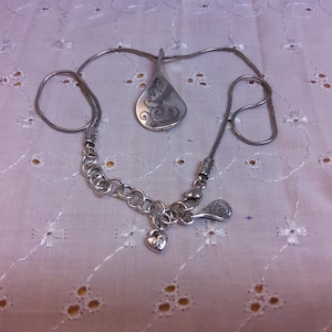 May include: A silver necklace with a pendant shaped like a stylized leaf with a swirl design. The necklace has a chain with a heart charm.