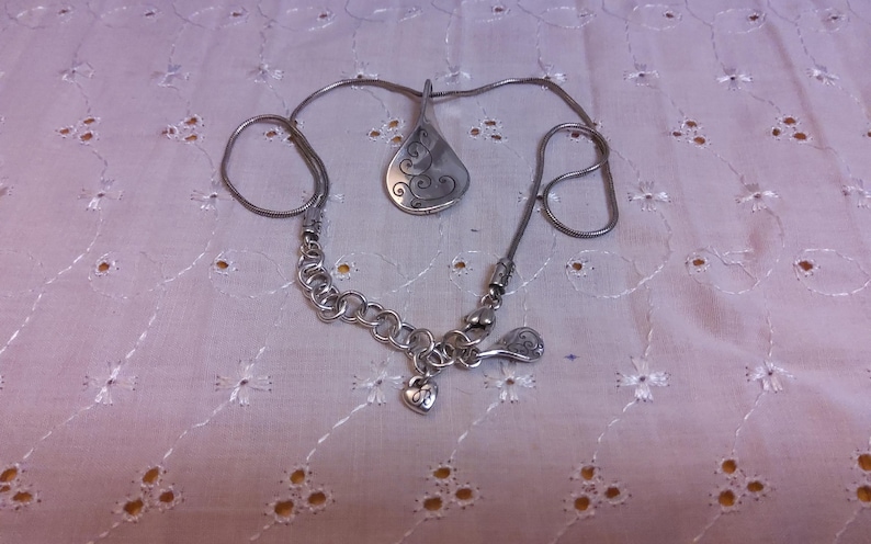 May include: A silver necklace with a pendant shaped like a stylized leaf with a swirl design. The necklace has a chain with a heart charm.