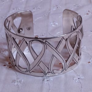 May include: Silver-toned cuff bracelet with an open design. The bracelet features a repeating pattern of intertwined ribbon shapes. The cuff has a hinged opening for easy wear and is displayed on a white, floral-patterned fabric.