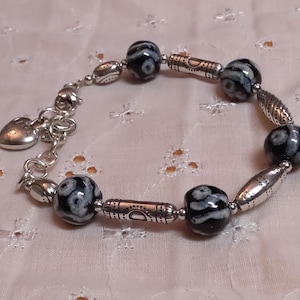 May include: A silver bracelet with black and white patterned beads and silver accents. The bracelet has a heart-shaped charm and is displayed on a white, patterned surface. The beads are round and cylindrical.