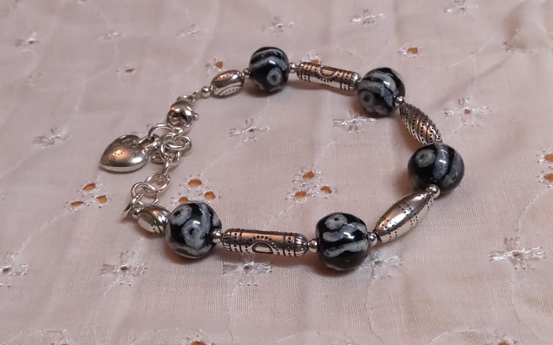 May include: A silver bracelet with black and white patterned beads and silver accents. The bracelet has a heart-shaped charm and is displayed on a white, patterned surface. The beads are round and cylindrical.
