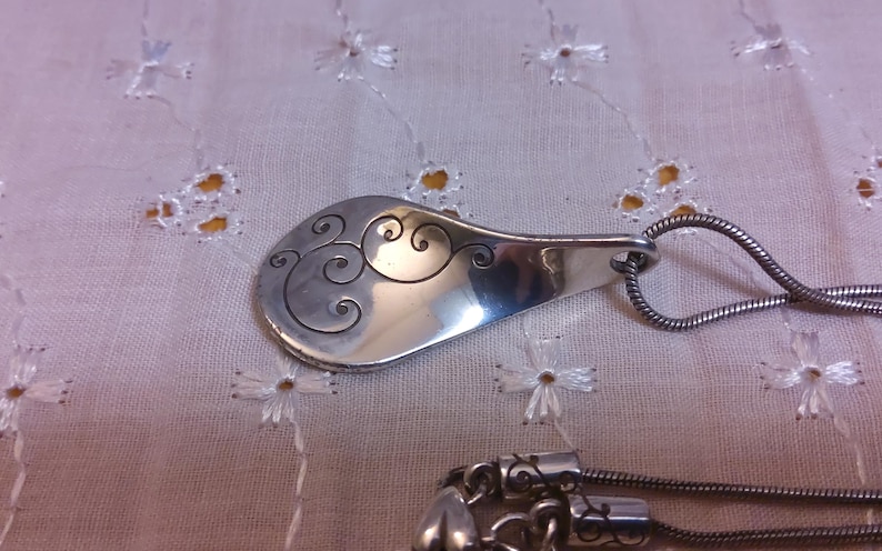 May include: A silver necklace with a pendant shaped like a spoon with a swirling design. The pendant is attached to a silver chain.