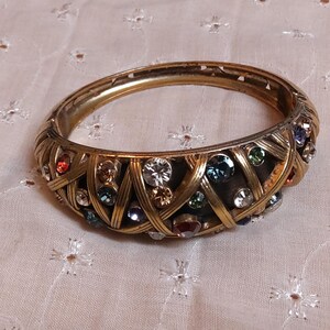 May include: Gold-toned hinged bangle bracelet with an intricate woven design. The bracelet is adorned with a variety of colorful gemstones, including clear, blue, red, and green stones. The bracelet is a piece of jewelry.