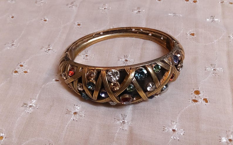 May include: Gold-toned hinged bangle bracelet with an intricate woven design. The bracelet is adorned with a variety of colorful gemstones, including clear, blue, red, and green stones. The bracelet is a piece of jewelry.
