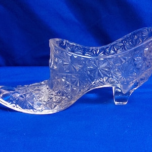 May include: Clear glass slipper-shaped decorative item. The shoe has a pointed toe, a small heel, and is decorated with a starburst pattern. The glass is transparent, allowing the blue background to show through.