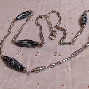 Op de afbeelding: Een zilverkleurige ketting met zwart-wit patronen kralen afgewisseld met diverse zilverkleurige metalen kralen. De ketting heeft een verstelbare ketting met een hartvormige bedel.