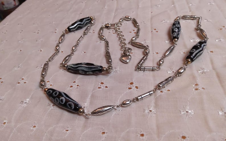 Op de afbeelding: Een zilverkleurige ketting met zwart-wit patronen kralen afgewisseld met diverse zilverkleurige metalen kralen. De ketting heeft een verstelbare ketting met een hartvormige bedel.