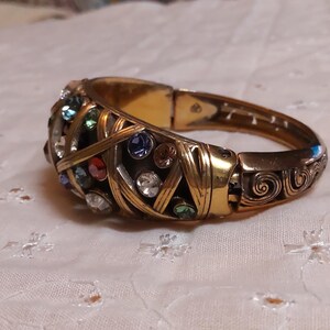 May include: Gold-toned hinged bracelet with an ornate design. The bracelet features a series of colorful gemstones, including red, blue, green, and clear crystals. The metalwork has a woven pattern and decorative swirls.