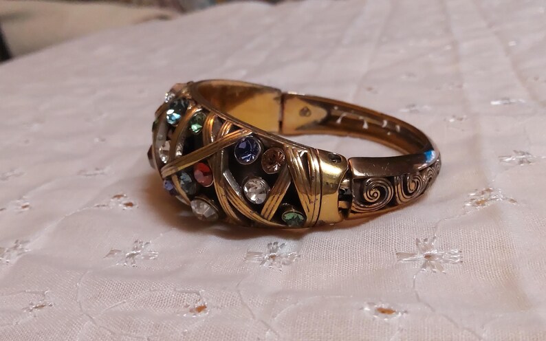 May include: Gold-toned hinged bracelet with an ornate design. The bracelet features a series of colorful gemstones, including red, blue, green, and clear crystals. The metalwork has a woven pattern and decorative swirls.