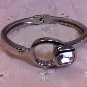 May include: A silver-toned hinged bracelet with a large, faceted, square-cut clear crystal. The bracelet has a simple, modern design with a clasp closure. The crystal is the focal point of the bracelet.