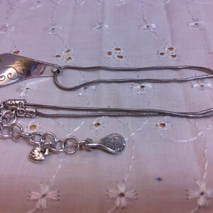 May include: A silver necklace with a pendant shaped like a teardrop with a swirl design. The necklace has a chain with a lobster clasp and a small heart charm.