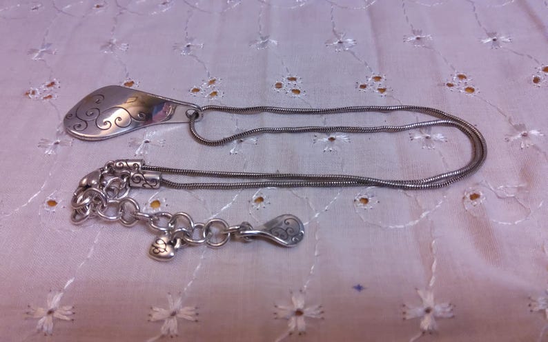 May include: A silver necklace with a pendant shaped like a teardrop with a swirl design. The necklace has a chain with a lobster clasp and a small heart charm.