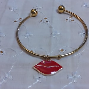 May include: Gold-tone bangle bracelet with a red lip charm. The charm has a gold outline and a small, clear rhinestone. The bracelet has a simple, elegant design, perfect for everyday wear or special occasions.
