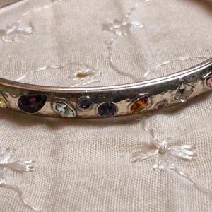 May include: Silver-toned bangle bracelet adorned with colorful gemstones. The bracelet features a variety of shapes and colors, including blue, purple, yellow, and pink stones. The gemstones are set in a hammered metal band, creating a textured appearance.