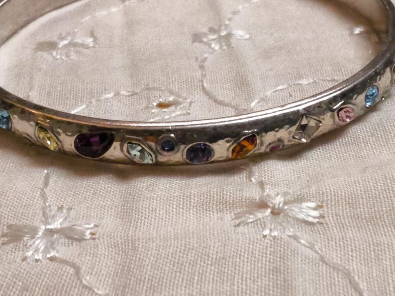 May include: Silver-toned bangle bracelet adorned with colorful gemstones. The bracelet features a variety of shapes and colors, including blue, purple, yellow, and pink stones. The gemstones are set in a hammered metal band, creating a textured appearance.