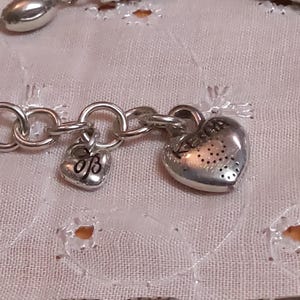 Op de afbeelding: Close-up van een zilverkleurige armband met hart- en bedelhangers. De armband heeft een schakelkettingontwerp met een kleine hartvormige bedel en een grotere hartvormige hanger met een gestippeld patroon.