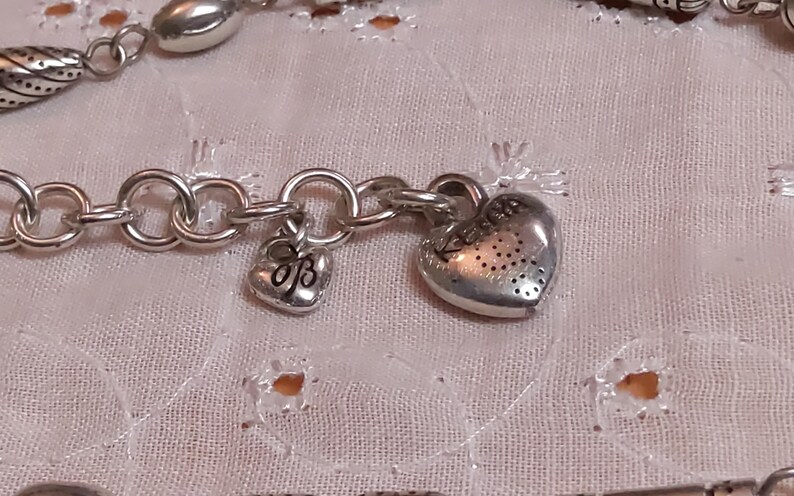 Op de afbeelding: Close-up van een zilverkleurige armband met hart- en bedelhangers. De armband heeft een schakelkettingontwerp met een kleine hartvormige bedel en een grotere hartvormige hanger met een gestippeld patroon.