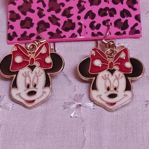 Pu&ograve; includere: Un paio di orecchini di Minnie Mouse. Ogni orecchino presenta un viso bianco, orecchie nere e un fiocco rosso a pois bianchi. Gli orecchini sono dorati e pendono da un gancio.