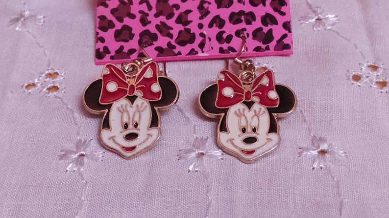 Pu&ograve; includere: Un paio di orecchini di Minnie Mouse. Ogni orecchino presenta un viso bianco, orecchie nere e un fiocco rosso a pois bianchi. Gli orecchini sono dorati e pendono da un gancio.