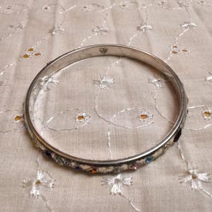 May include: Silver-toned bangle bracelet with colorful gemstones. The bracelet features a row of small, multi-colored stones set along the outer edge. A small heart design is visible on the bracelet. The bracelet is resting on a cream-colored, embroidered fabric.