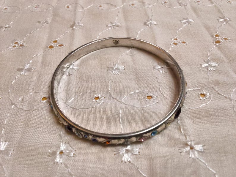 May include: Silver-toned bangle bracelet with colorful gemstones. The bracelet features a row of small, multi-colored stones set along the outer edge. A small heart design is visible on the bracelet. The bracelet is resting on a cream-colored, embroidered fabric.