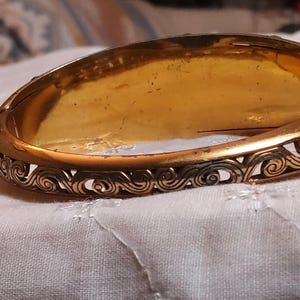 May include: A vintage gold-toned hinged bangle bracelet. The bracelet features an ornate, openwork design along the edges with a repeating swirl pattern. The interior has a warm, golden hue, suggesting age and wear.