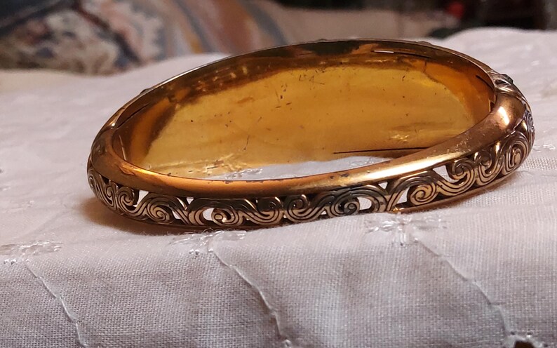 May include: A vintage gold-toned hinged bangle bracelet. The bracelet features an ornate, openwork design along the edges with a repeating swirl pattern. The interior has a warm, golden hue, suggesting age and wear.