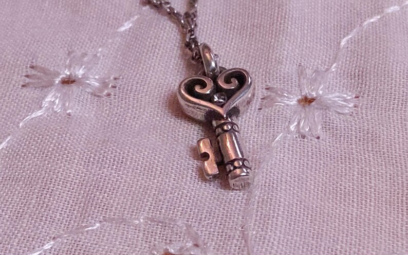 Op de afbeelding: Zilverkleurige sleutelhangerketting met een hartvormige bovenkant en een kleine edelsteen. De sleutel hangt aan een delicate ketting, tegen een lichtroze stof met geborduurde bloemendetails. Een romantisch sieraad.