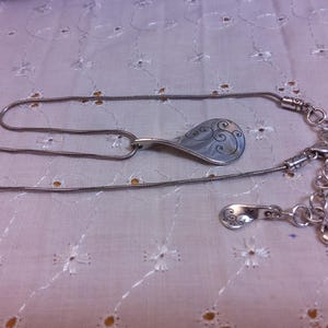 May include: A silver chain necklace with a silver pendant shaped like a teardrop with a swirl design. The necklace has a lobster clasp and a small silver charm.