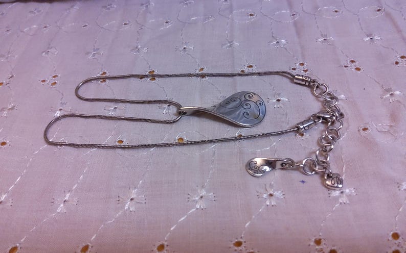 May include: A silver chain necklace with a silver pendant shaped like a teardrop with a swirl design. The necklace has a lobster clasp and a small silver charm.
