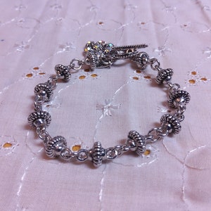 May include: A silver bracelet with a charm made of rhinestones. The bracelet is made of small silver beads connected by a chain. The charm is attached to the bracelet with a silver loop.
