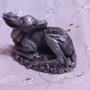 May include: A small, detailed pewter figurine of a resting rabbit. The rabbit is curled up on a bed of foliage, with its ears perked up. The figurine has a metallic gray finish and is set against a light pink background.