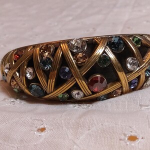 May include: A gold-toned bangle bracelet with a woven design, encrusted with a variety of colorful gemstones. The gemstones include clear, blue, red, and green stones. The bracelet is resting on a white, textured surface.
