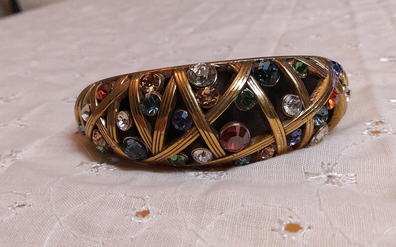 May include: A gold-toned bangle bracelet with a woven design, encrusted with a variety of colorful gemstones. The gemstones include clear, blue, red, and green stones. The bracelet is resting on a white, textured surface.