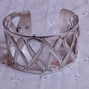May include: Silver-toned cuff bracelet with an open design. The bracelet features an intricate pattern of interwoven ribbon-like shapes. The cuff is wide and has a polished finish, suitable for everyday wear or special occasions.