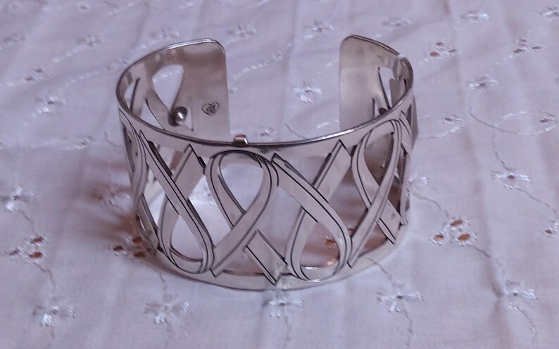 May include: Silver-toned cuff bracelet with an open design. The bracelet features an intricate pattern of interwoven ribbon-like shapes. The cuff is wide and has a polished finish, suitable for everyday wear or special occasions.
