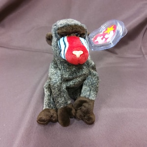 May include: A brown and gray stuffed monkey with a red face and a blue and white stripe across its nose. The monkey is sitting with its legs crossed and has a Ty Beanie Baby tag attached to its ear.