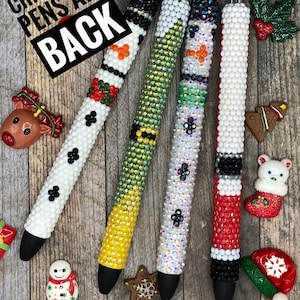 Puede incluir: Cuatro bolígrafos con temática navideña con pedrería. Los bolígrafos están decorados con un muñeco de nieve, un reno, un Papá Noel y un árbol de Navidad. El texto "Christmas Pens Are Back" es visible en la imagen.