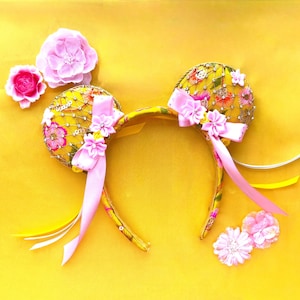 May include: A yellow headband featuring two circular ears covered in yellow fabric with a floral pattern, intricate gold embroidery, and sequins. The ears are decorated with pink fabric flowers, light pink satin ribbons, and dangling yellow ribbons.